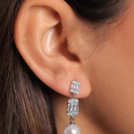 A close-up lifestyle photograph of a model wearing a geometric, vertical drop earring finished in a bright silver color, featuring a tiered stack of two crystal-encrusted rectangular frames suspending a smooth, luminous white teardrop pearl