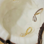 A pair of elegant gold-tone huggie hoop earrings, each featuring a shimmering pavé-set band and a delicate, dangling pear-cut crystal drop, displayed inside a coconut shell